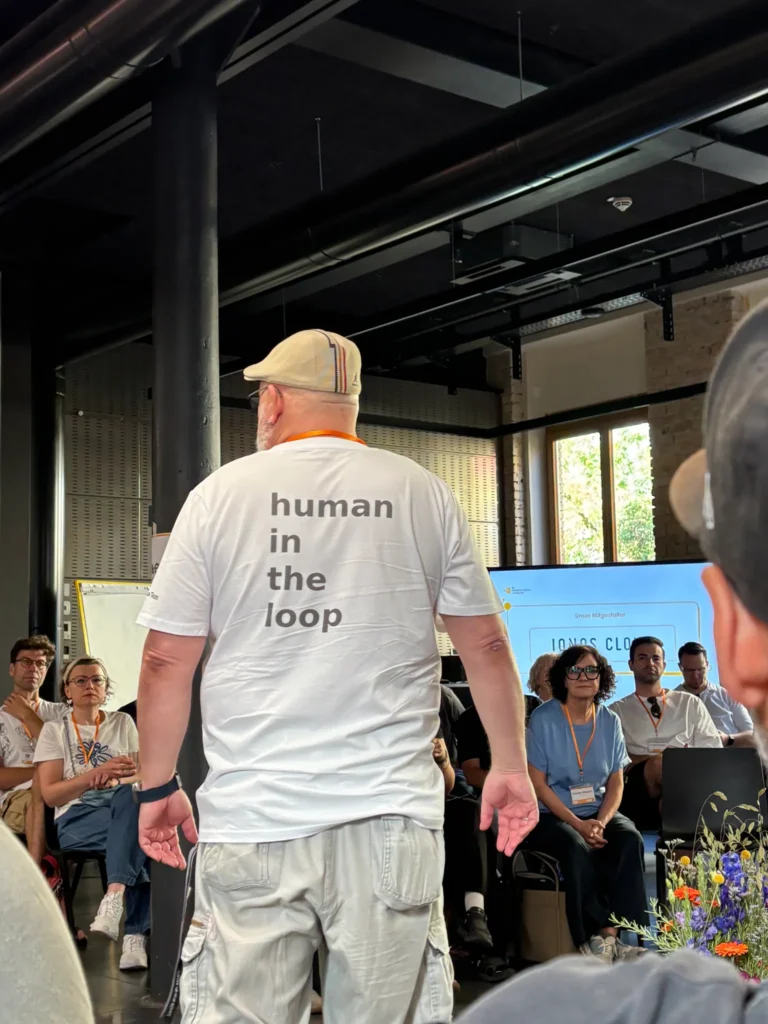 Eine Person mit einem T-Shirt mit der Aufschrift „human in the loop“ steht vor einer sitzenden Gruppe in einem Seminarraum.