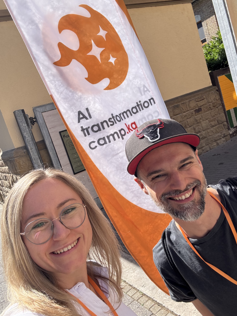 Benjamin Burkardt und Elisa Heinzelmann von ideenhunger stehen vor der Fahne des AI Transformation Camp und machen ein Selfie.